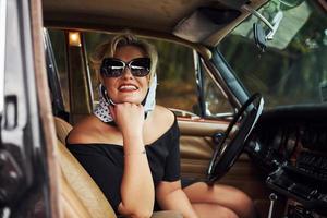 mujer rubia con gafas de sol y vestido negro se sienta en un viejo coche clásico vintage foto