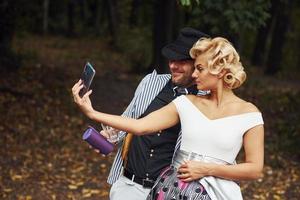 Making a selfie. Beautiful couple in old fashioned wear near retro car with forest at background photo