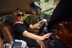 mujer rubia con gafas de sol y vestido negro se sienta en un viejo coche clásico vintage foto