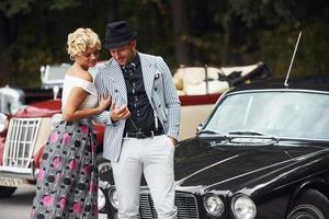 Beautiful couple in old fashioned wear near retro car with forest at background photo