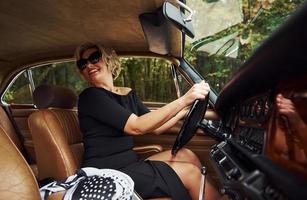 Blonde woman in sunglasses and in black dress sits in old vintage classic car photo