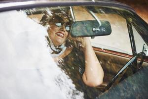 Blonde woman in sunglasses and in black dress sits in old vintage classic car photo