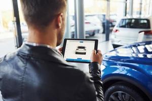 Man stands inside car salon with tablet in hands and looks at the vehicle picture