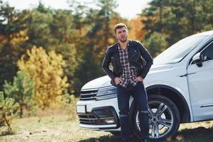 Man in black leather jacket stands near his parked white car outdoors and waits for help photo