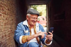 senior elegante con ropa de moda y gafas se sienta en la silla y usa el teléfono foto