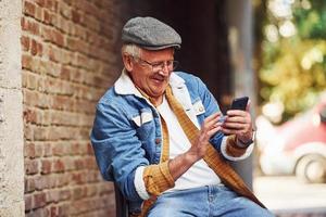 Stylish senior in fashionable clothes and in glasses sits on the chair and uses phone photo