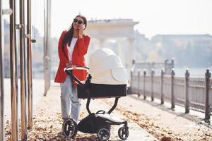 Mother in red coat have a walk with her kid in the pram in the park at autumn time photo