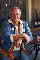 Stylish senior in fashionable clothes and in glasses sits in the cafe with drink and laptop on the table photo