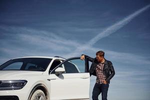 Man in black leather jacket stands near his parked white car outdoors against blue sky photo