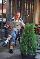 Stylish senior in fashionable clothes and in glasses sits in the cafe and reads newspaper photo