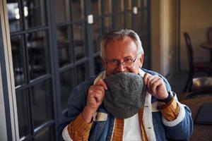 Stylish senior in fashionable clothes and in glasses sits in the cafe and holds hat photo