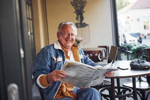 senior con estilo en ropa de moda y con gafas se sienta en el café y lee el periódico foto