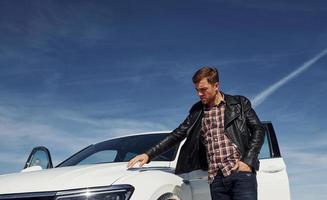 Man in black leather jacket stands near his parked white car outdoors against blue sky photo