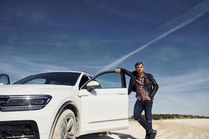 Man in black leather jacket stands near his parked white car outdoors against blue sky photo