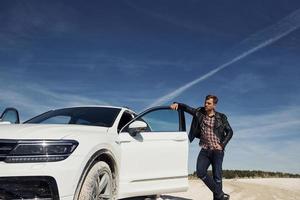 Man in black leather jacket stands near his parked white car outdoors against blue sky photo