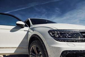 Front view of white modern car parked outdoors against blue sky at background and with dirt on headlights photo