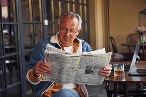 senior con estilo en ropa de moda y con gafas se sienta en el café y lee el periódico foto