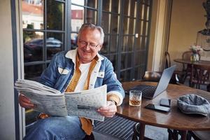 Stylish senior in fashionable clothes and in glasses sits in the cafe and reads newspaper photo
