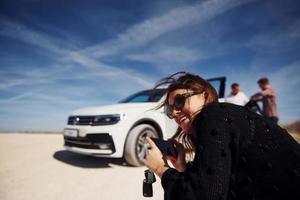 Girl with phone in hands smiling and sits against white car and people at background photo