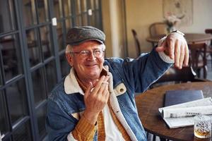 Stylish senior in fashionable clothes and in glasses sits in the cafe with cigarette and talks photo