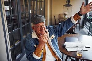 Stylish senior in fashionable clothes and in glasses sits in the cafe with cigarette and talks photo