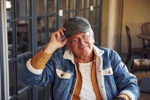 Stylish senior in fashionable clothes and in glasses sits in the cafe and holds hat photo