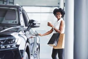 joven afroamericana con anteojos se para en un salón de autos cerca del vehículo con un paquete en las manos foto