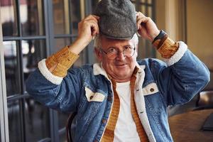 senior con estilo en ropa de moda y con gafas se sienta en el café y sostiene el sombrero foto