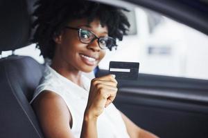 tiene tarjeta de crédito. una joven afroamericana se sienta dentro de un auto nuevo y moderno foto