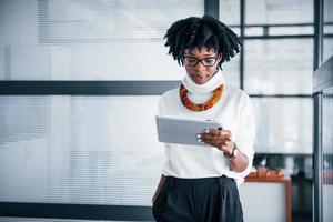 Young african american woman in glasses stands indoors in the office with tablet in hands photo