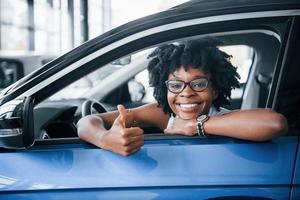 muestra el pulgar hacia arriba. una joven afroamericana se sienta dentro de un auto nuevo y moderno foto