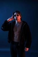 hermoso hombre con gafas y ropa negra está en el estudio con luces de neón azul foto