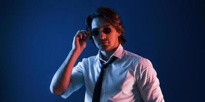 un hombre hermoso con ropa formal y gafas está en el estudio con luces de neón azul foto