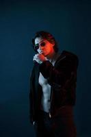 Beautiful man in glasses and black clothes is in the studio with blue neon lighting photo