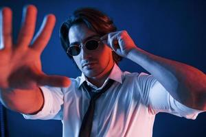Beautiful man in glasses is in the studio with blue neon lighting holds hand in front of him photo