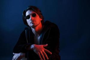 Beautiful man in glasses and black clothes is in the studio with blue neon lighting photo