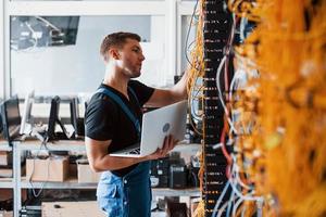 Young man in uniform and with laptop works with internet equipment and wires in server room photo