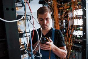 Young man in uniform with measuring device works with internet equipment and wires in server room photo