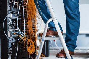 Man in uniform stands on the ladder and works with internet equipment and wires in server room photo