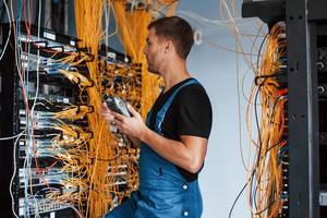 Young man in uniform with measuring device works with internet equipment and wires in server room photo