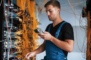 Young man in uniform with measuring device works with internet equipment and wires in server room photo