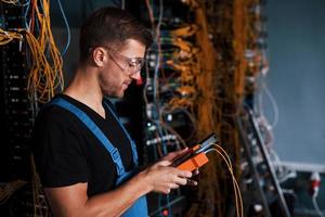 Young man in uniform with measuring device works with internet equipment and wires in server room photo