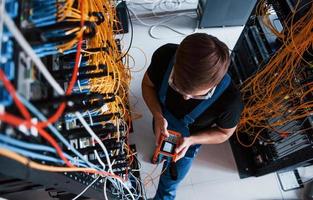Top view of young man in uniform with measuring device that works with internet equipment and wires in server room photo