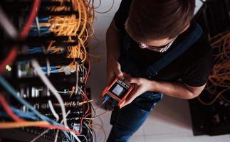 Top view of young man in uniform with measuring device that works with internet equipment and wires in server room photo