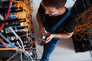 Top view of young man in uniform with measuring device that works with internet equipment and wires in server room photo