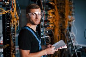 Young man in uniform with notepad in hands works with internet equipment and wires in server room photo