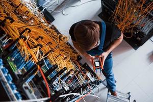 Top view of young man in uniform with measuring device that works with internet equipment and wires in server room photo