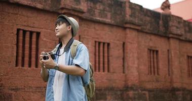 glücklicher asiatischer Reisender mit Hut macht ein Foto und besucht den alten Tempel. lächelnder junger Mann, der Kamera steht und hält. urlaubs-, reise- und hobbykonzept. video
