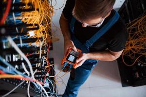 Top view of young man in uniform with measuring device that works with internet equipment and wires in server room photo