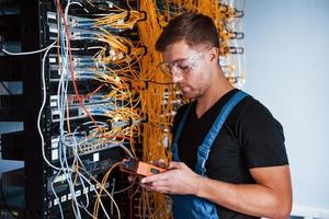 Young man in uniform with measuring device works with internet equipment and wires in server room photo